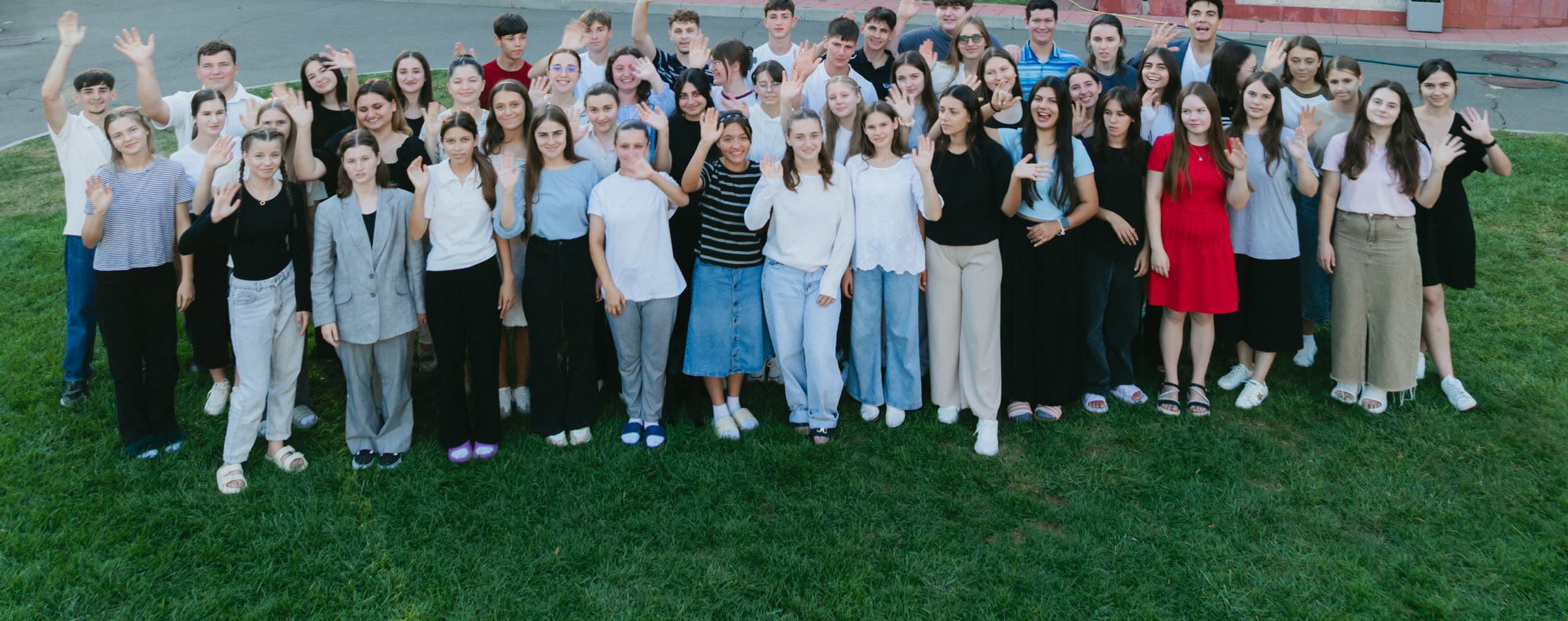 Around fifty children and teenagers standing together on a lawn, smiling and waving at the camera in evening light.